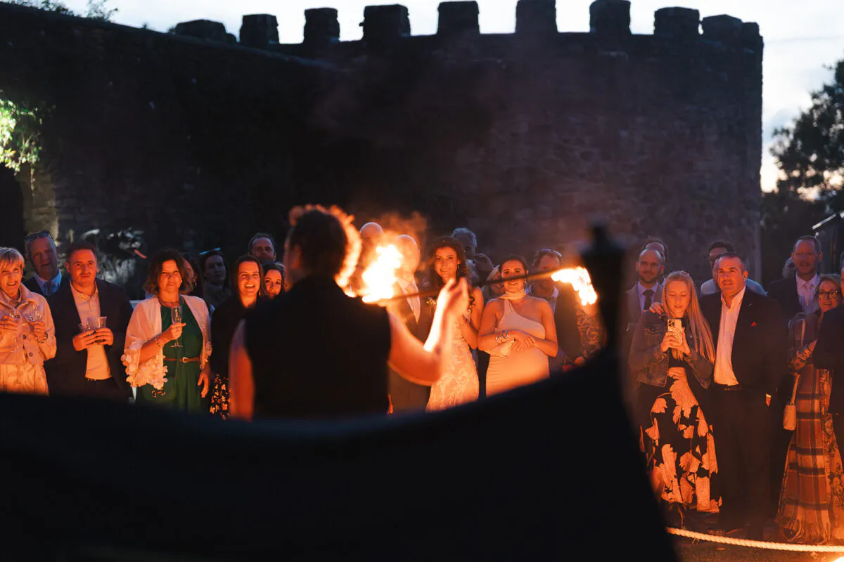 Wedding guests watch a dramatic fire performer put on an evening show against the backdrop of a historic stone castle at dusk.