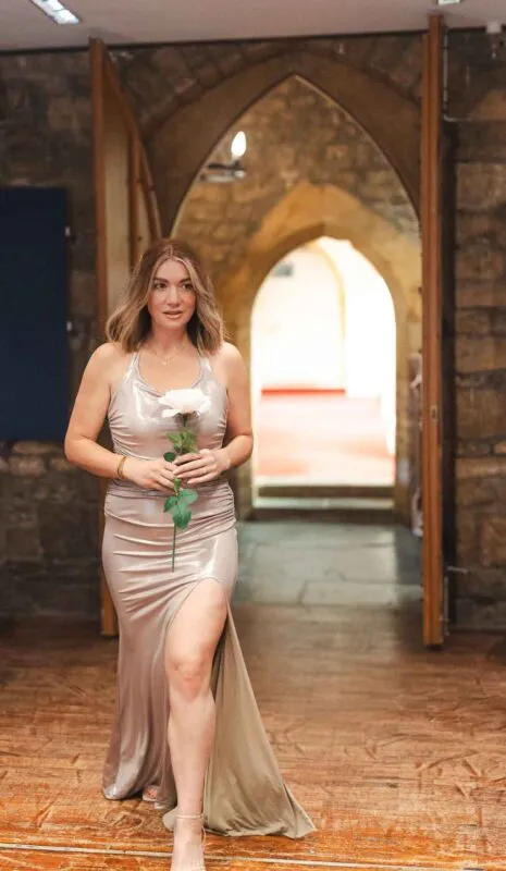 A smiling bride holding a single rose walks forward through a stone archway in a venue with exposed brick and wooden floors.