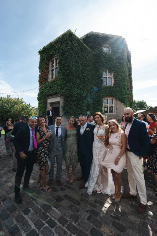 The bride and groom pose with wedding guests in a cobbled courtyard outside a historic stone building covered in lush green ivy under bright sunlight.