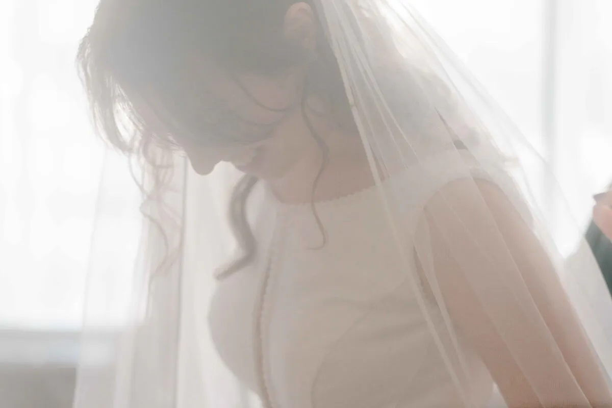 A beautiful bride smiles softly in a bright room, framed gently by a sheer veil during wedding morning preparations.