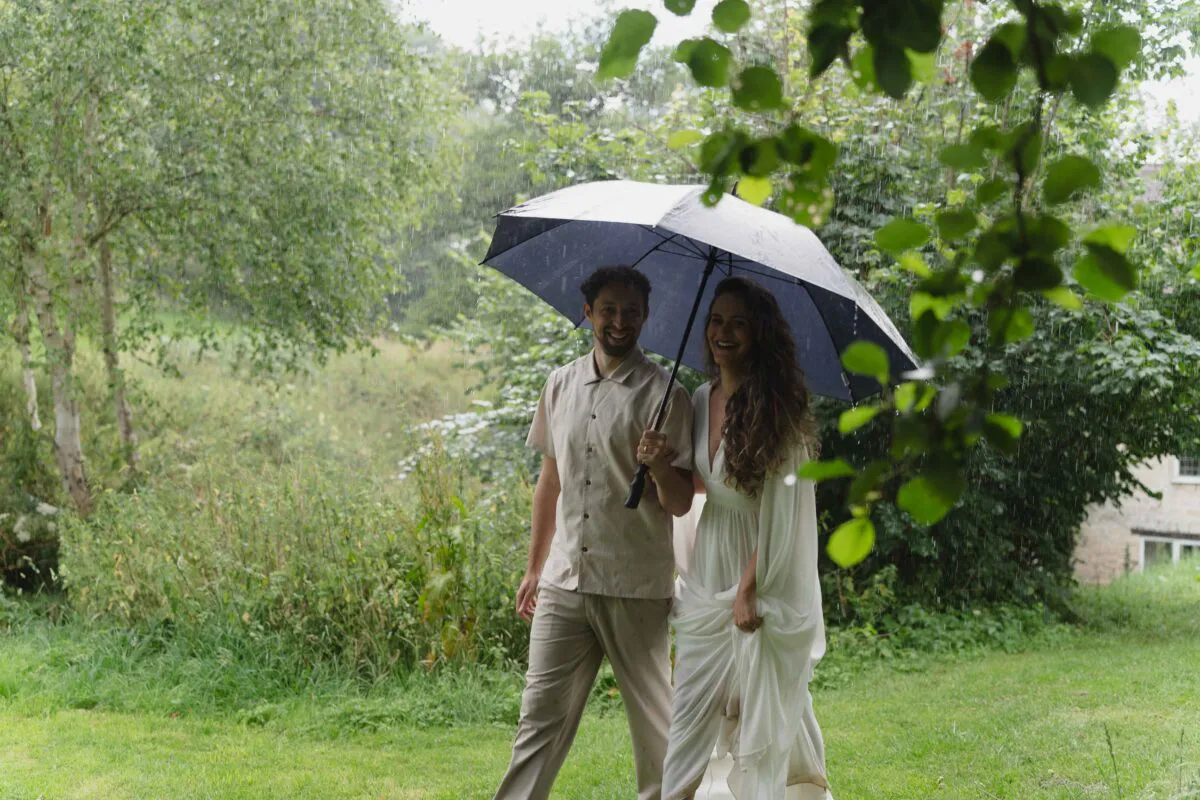 A smiling bride and groom walk through a lush green woodland holding an umbrella together amidst falling rain.