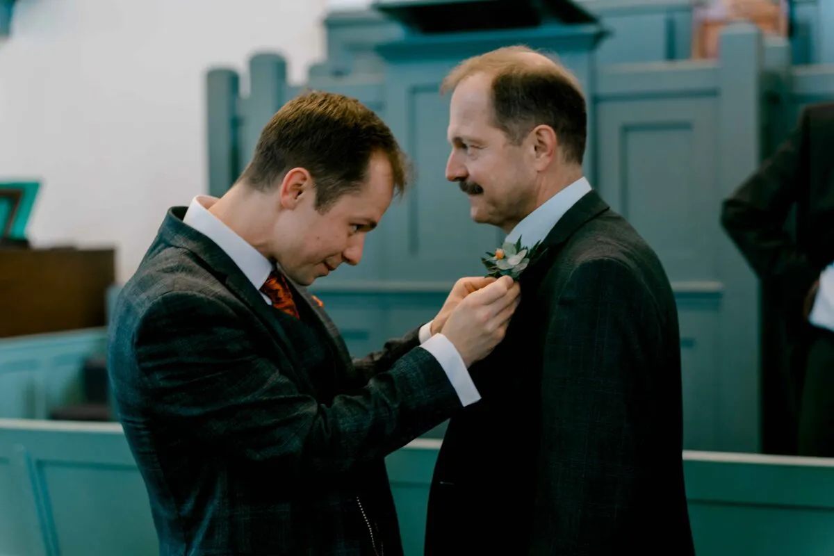 A groom carefully pins a boutonniere onto another man's lapel inside a church before the ceremony.