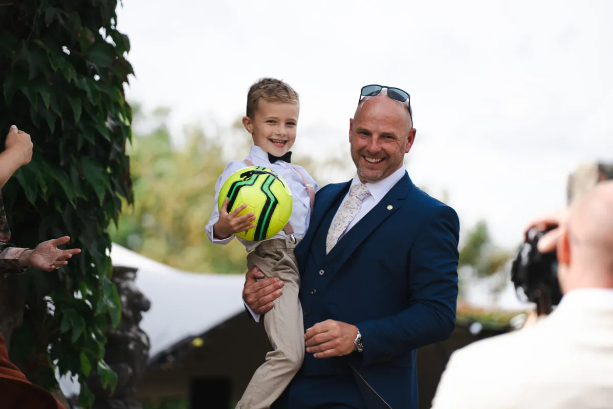 A smiling man in a navy three-piece suit holds a young page boy who is clutching a bright yellow and black football outdoors near an ivy-covered wall.