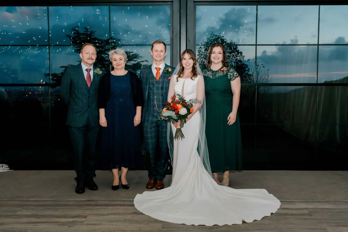 A bridal party and family members pose for a group portrait indoors against a large grid-panelled window during twilight.