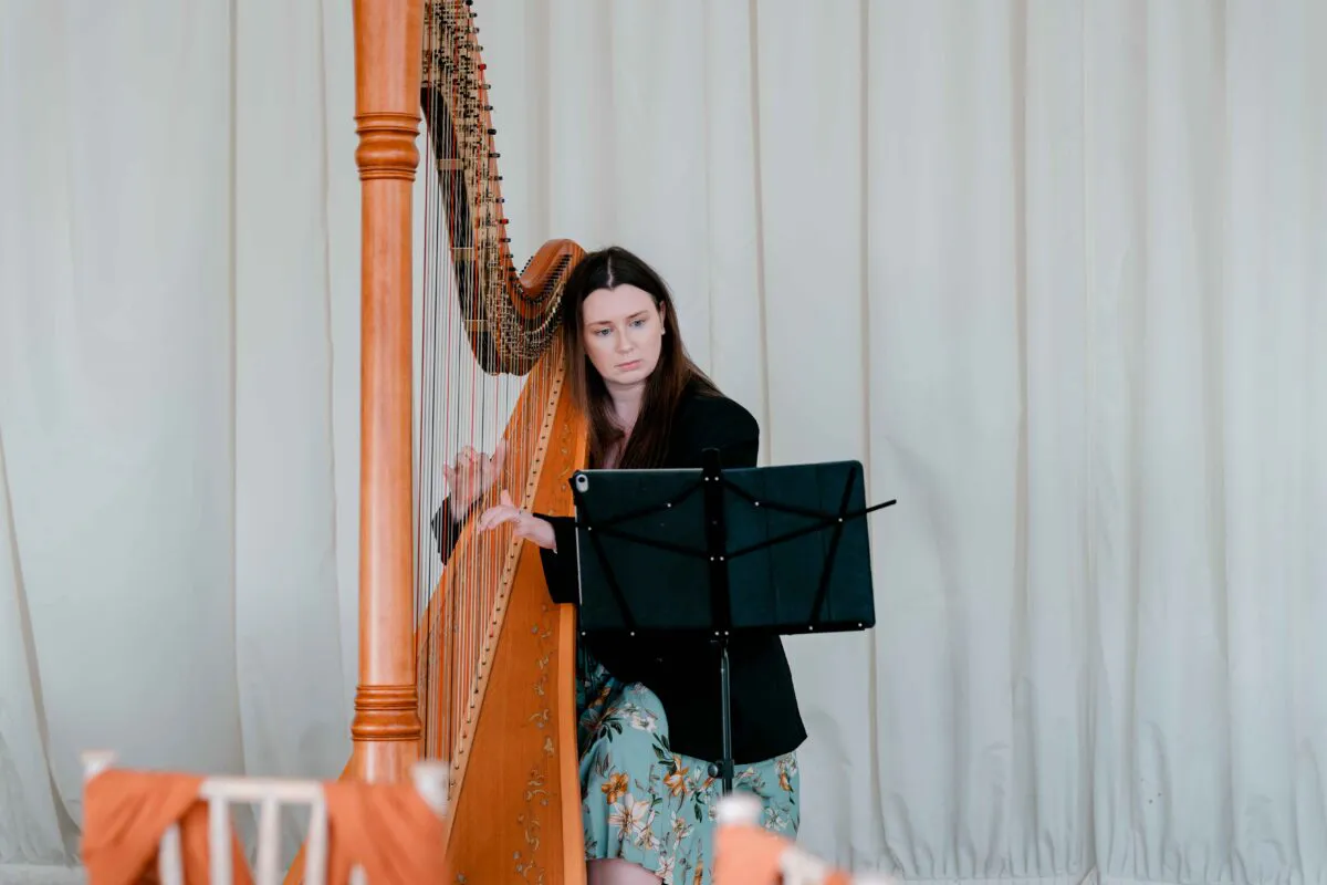 A professional harpist performs on a wooden harp during a wedding ceremony in front of draped white fabric.