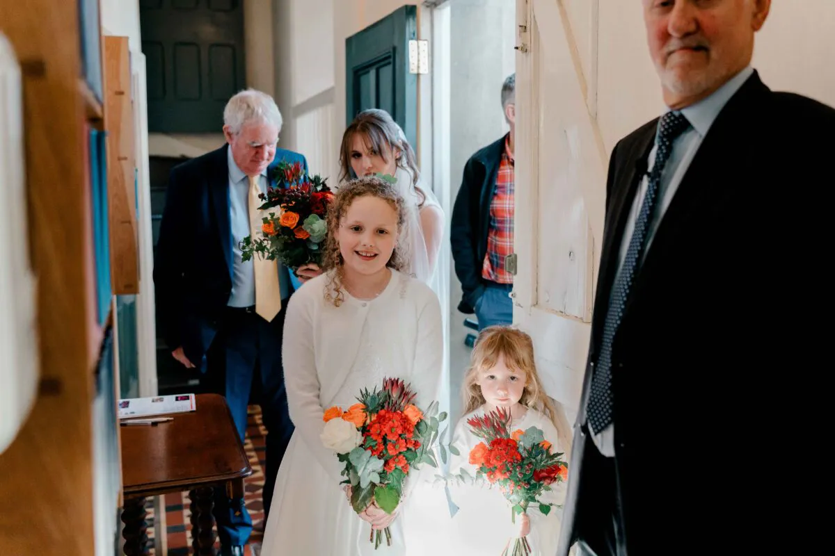 A smiling flower girl holding a colourful bouquet of orange and white flowers stands in a doorway as the bridal party prepares to enter the ceremony.