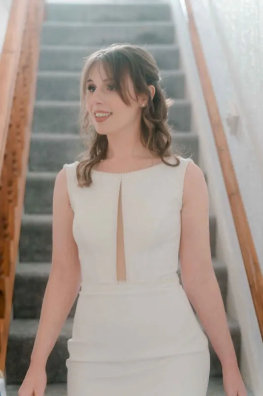 A glowing bride poses indoors near a carpeted staircase wearing a sleek white crepe dress with a keyhole neckline and half-up cascading waves.