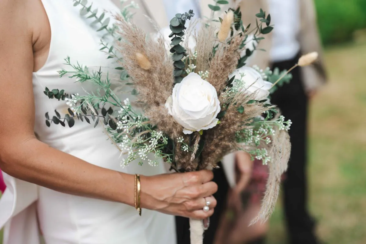 A bride holds a textured, bohemian-inspired hand-tied bouquet featuring white roses, pampas grass, eucalyptus, and dried grasses against a simple white gown.
