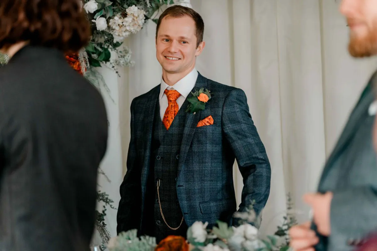A smiling groom wearing a checked three-piece suit with an orange patterned tie and matching pocket square stands at the altar during his wedding ceremony.