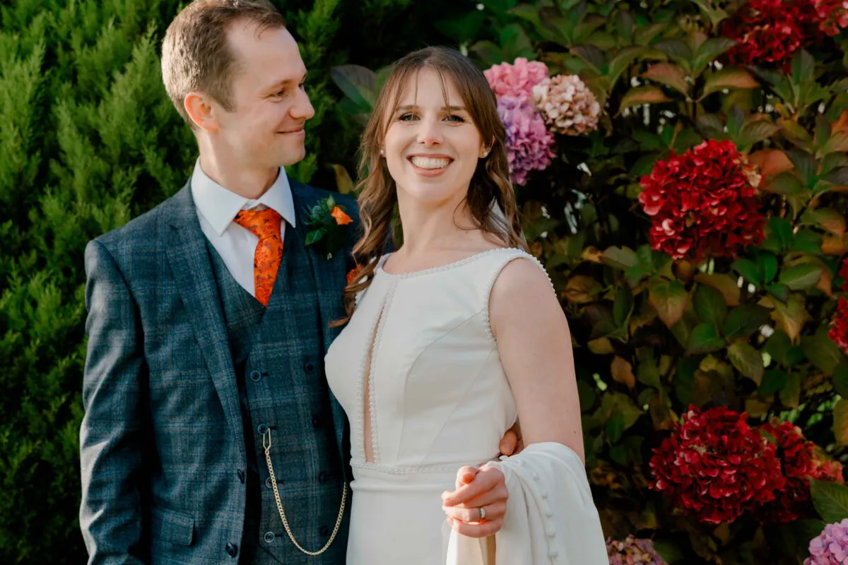 A smiling bride and groom pose together outdoors against a backdrop of lush green foliage and vibrant red and pink hydrangeas.