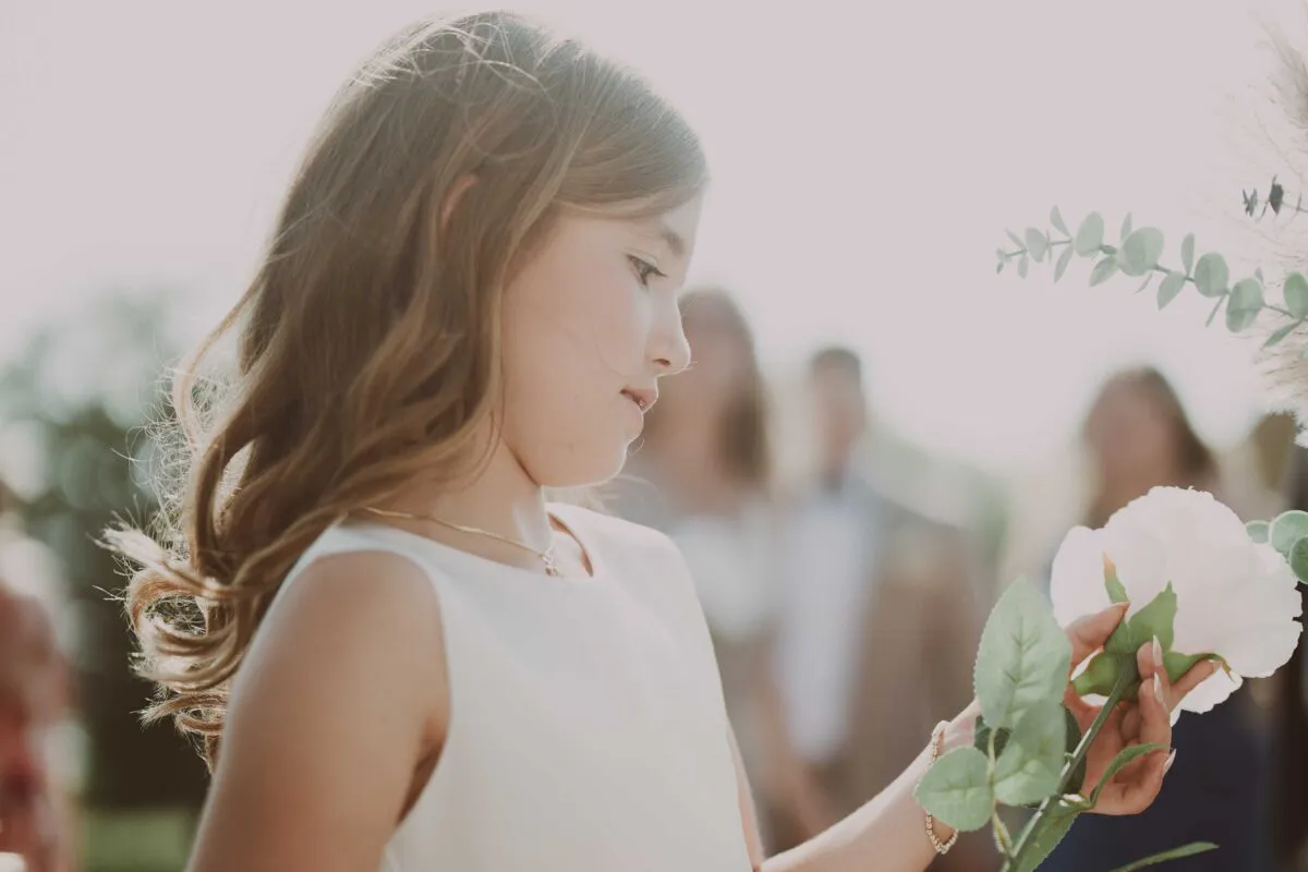 A young flower girl in a white dress gently holds a white rose, illuminated by soft backlit natural light with a blurred background.