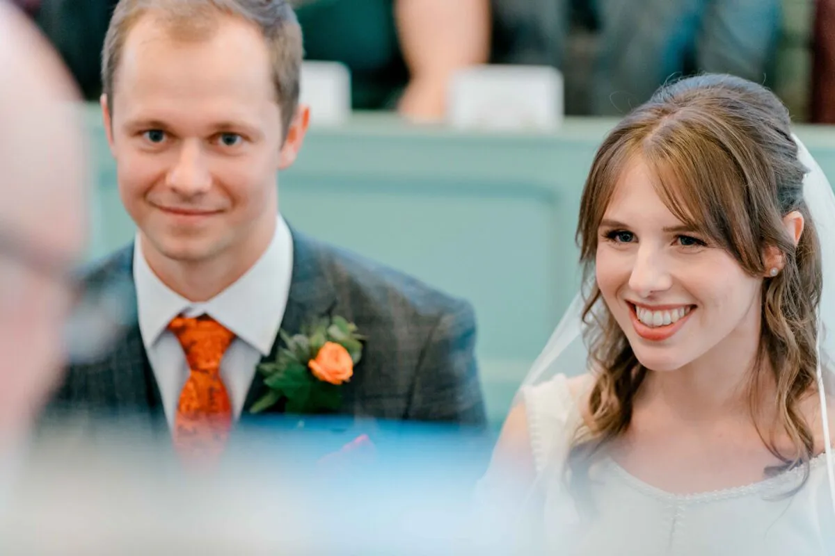 A smiling bride and groom sitting together during their wedding ceremony, with the groom wearing a grey suit and a bright orange tie.