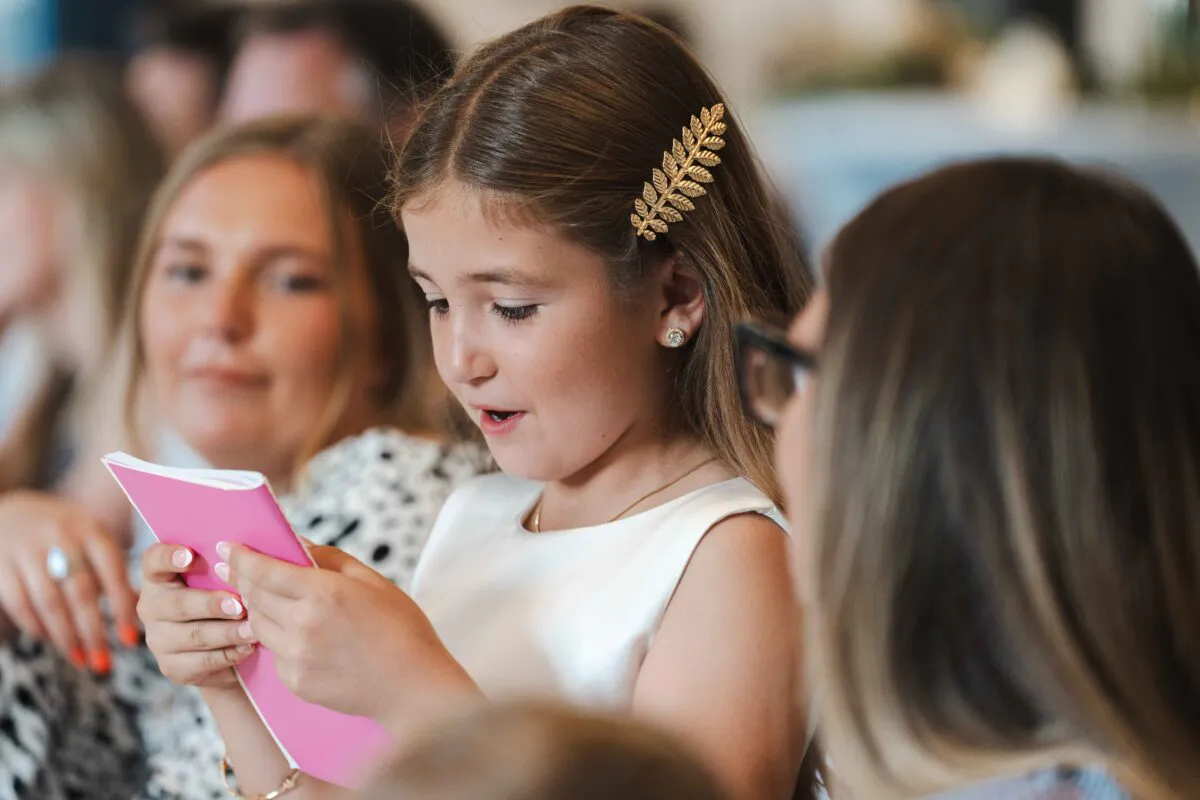 A young flower girl wearing a gold leaf hair accessory holds a small pink booklet while participating in the wedding ceremony.