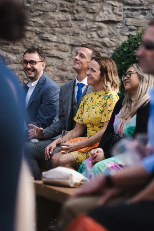 Wedding guests smile and enjoy themselves during an outdoor drinks reception in front of a rustic stone wall.