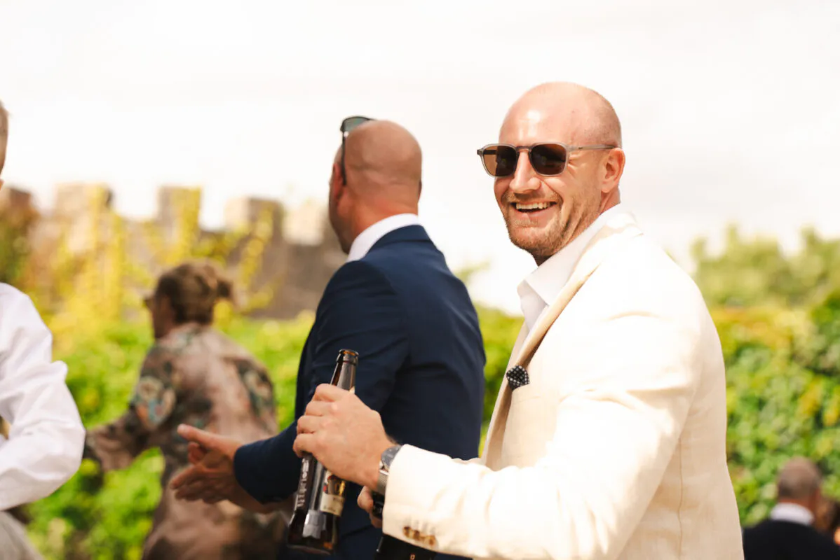 A smiling guest wearing a cream suit and sunglasses looks back toward the camera during an outdoor wedding drinks reception.