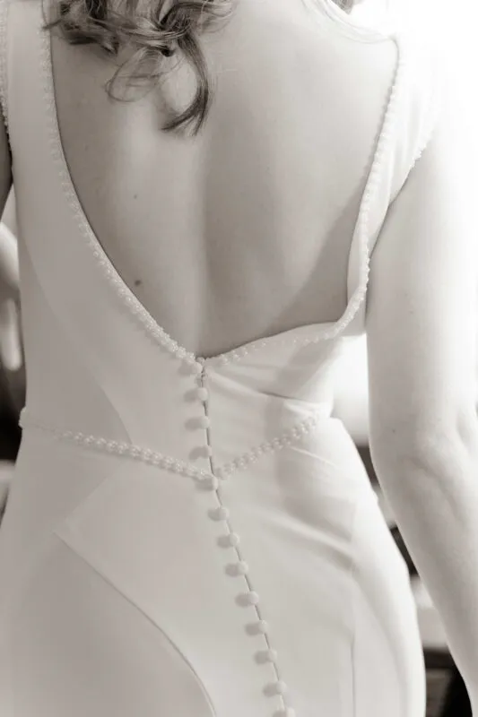 A back view of a bride wearing a white crepe wedding gown featuring pearl trim along the low scoop neckline and a line of delicate fabric buttons down the back.