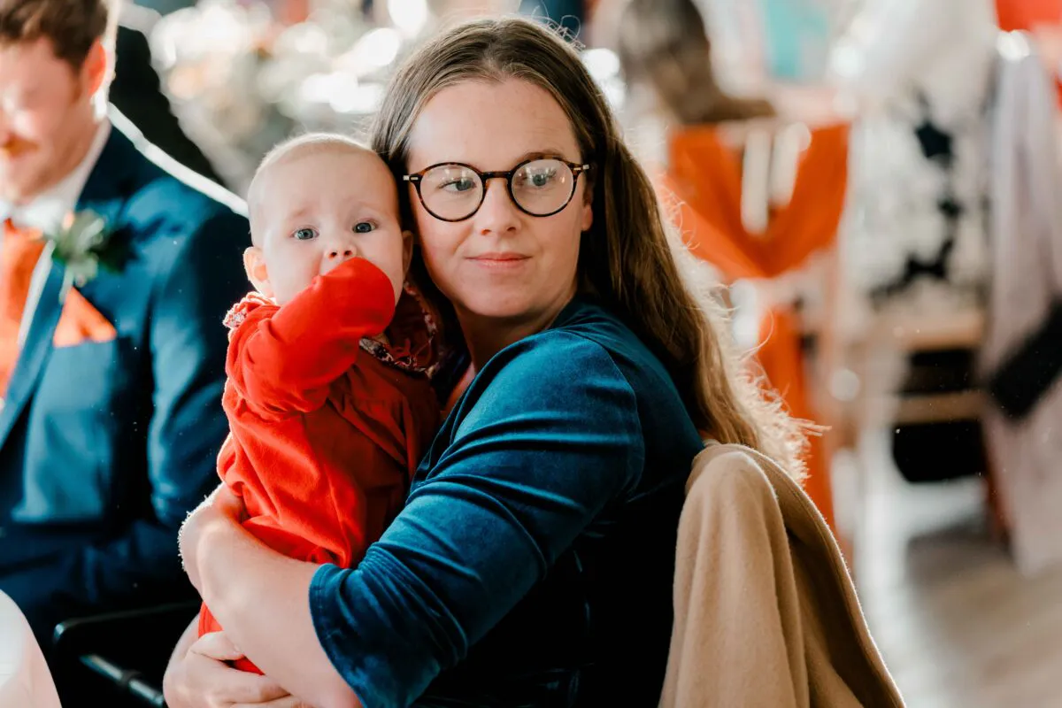 A guest wearing glasses and a dark blue top gently holds a baby dressed in a bright red outfit during a wedding celebration.