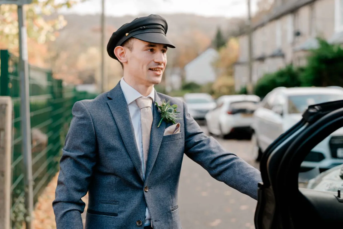 A smiling groom wearing a textured grey suit and a stylish black cap opens the door of a car on a residential street.