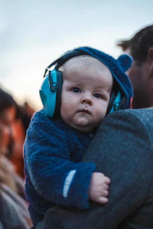 A baby wearing bright blue ear defenders is held safely over an adult's shoulder during a wedding celebration.