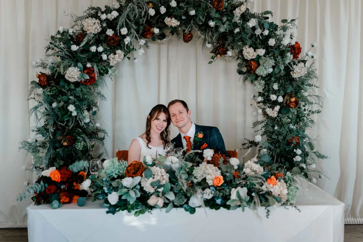 A smiling bride and groom sit behind a flower-adorned top table featuring a large circular floral arch with white and burnt orange blooms against a draped fabric backdrop.