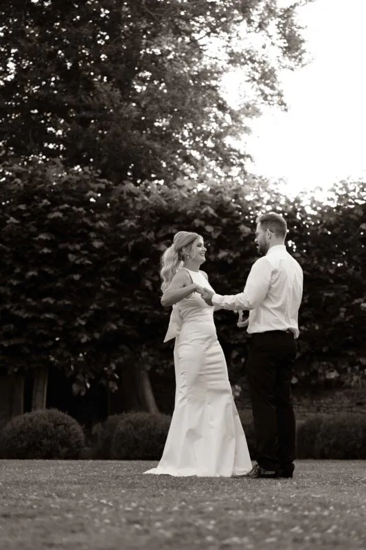 A newlywed couple shares a joyful moment dancing together outdoors in a manicured garden during golden hour, captured in a timeless black and white edit.