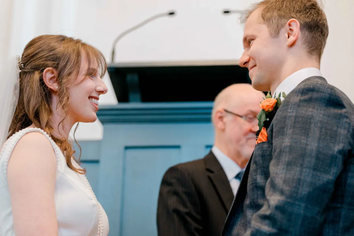 A smiling bride and groom gaze lovingly at each other during their wedding ceremony while an officiant looks on in the background.