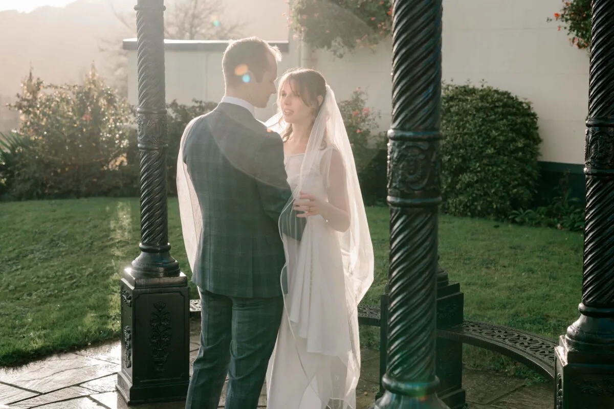 A newlywed couple embrace tenderly inside a wrought iron bandstand during golden hour, framed by dramatic lighting and warm sun flare.