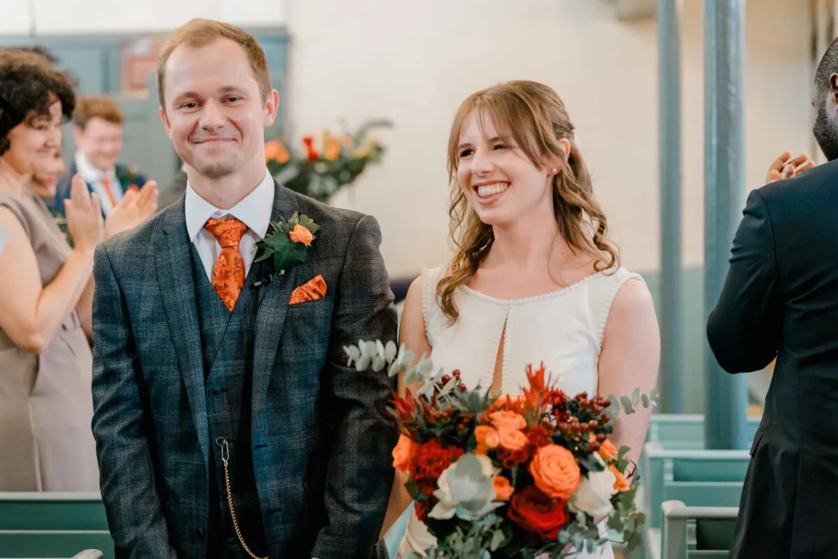 A smiling bride holding a vibrant autumn-toned bouquet and a groom in a charcoal tweed suit stand happily during their wedding ceremony.