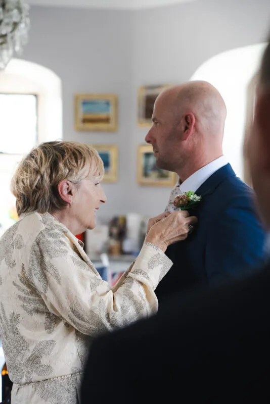 An older woman smiling warmly as she pins a small floral buttonhole onto the lapel of a groom in a navy suit.