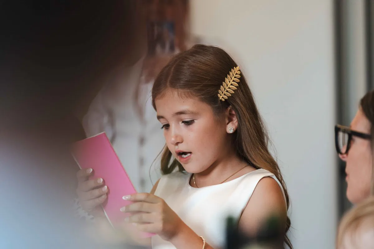 A young flower girl with a golden leaf hair pin looks down intently at a small pink booklet during the wedding morning preparations.