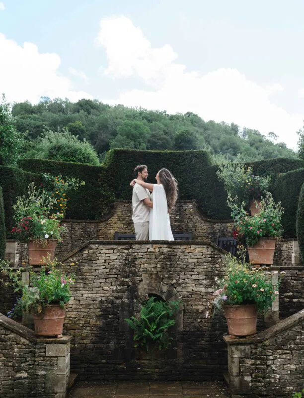A newlywed couple embraces lovingly on a stone bridge framed by potted plants and manicured hedges in a lush English country garden.