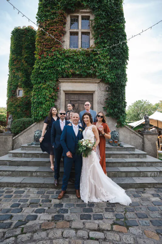 The bride and groom pose with family members on the stone steps outside an ivy-clad stone castle tower, holding a hand-tied bouquet.