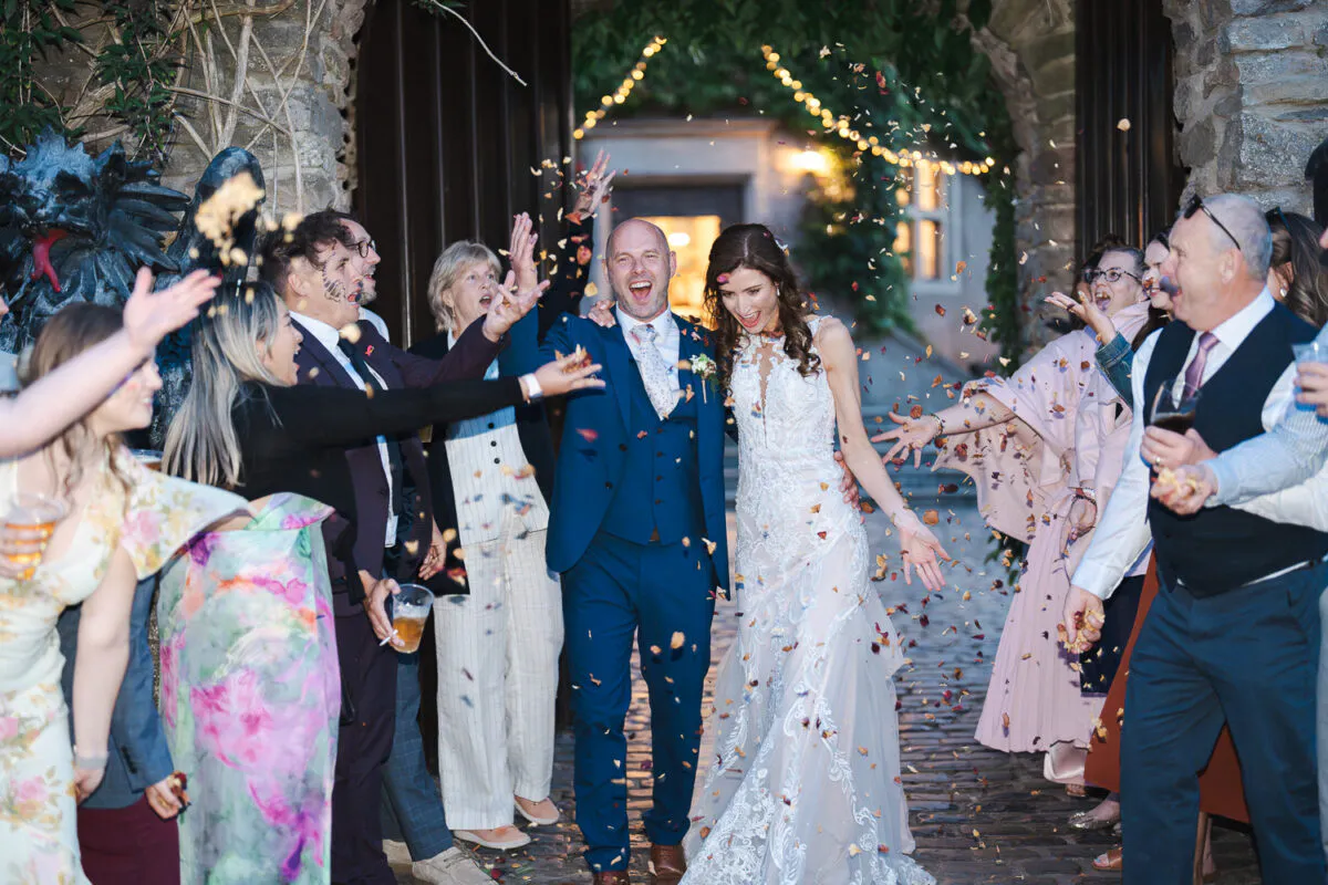 Newlyweds joyfully exit through a stone archway while cheering wedding guests shower them with a cascade of colorful confetti.