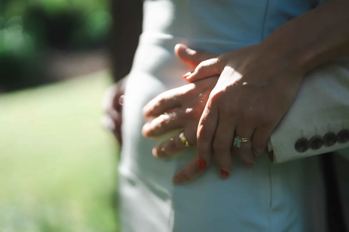 A close-up shot of a couple embracing with wedding rings and engagement ring visible on their hands against a softly blurred background.