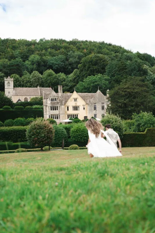 A newlywed couple runs joyfully across a sprawling green lawn towards a grand stone manor house nestled against a lush forested hillside.