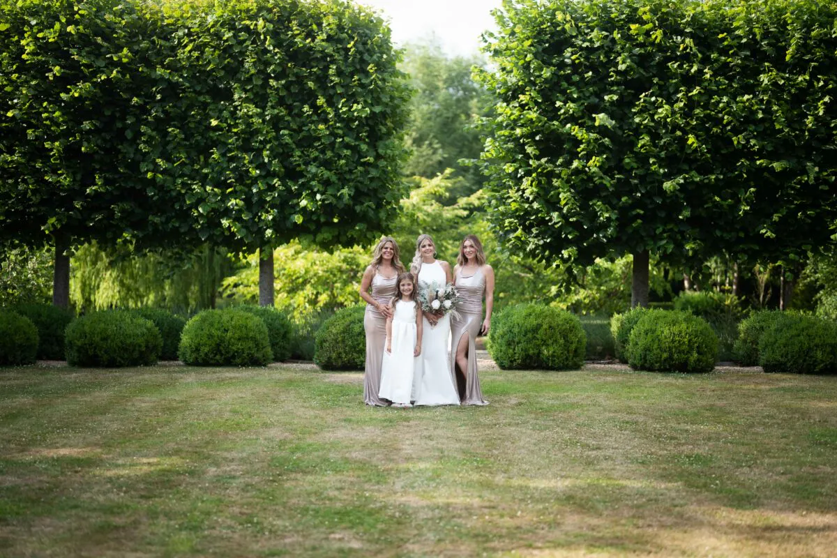 A beautiful bride stands in a manicured garden holding a bouquet, flanked by bridesmaids in satin dresses and a smiling flower girl.