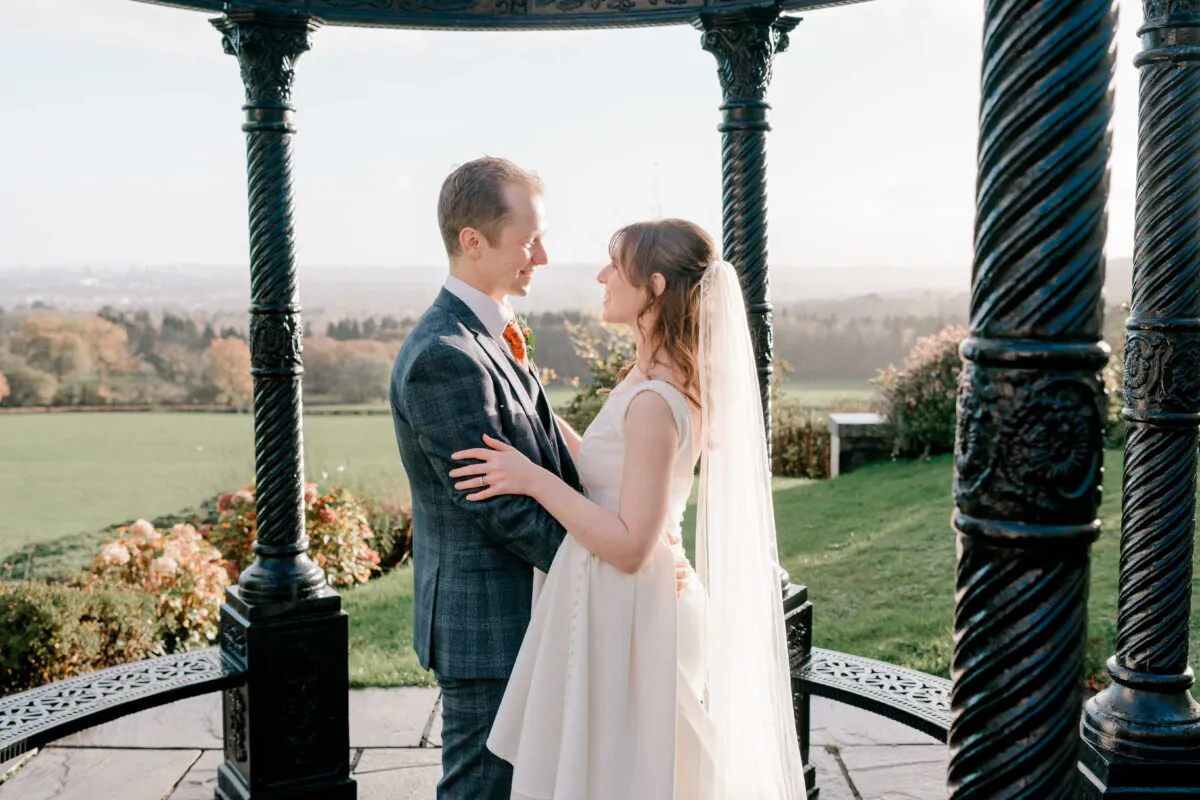 A bride and groom share a tender moment framed by an ornate black metal bandstand overlooking scenic rolling hills.