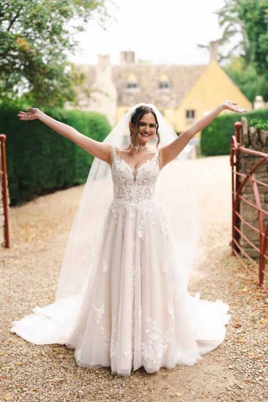 A joyful solo bride in an a-line lace wedding dress and chapel veil stands in a cobbled courtyard with arms outstretched, laughing in front of a country house.