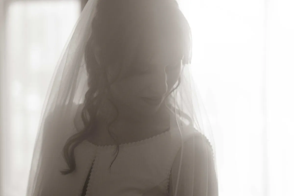 A close-up black and white portrait of a bride looking down softly with a veil framing her face against a bright window.