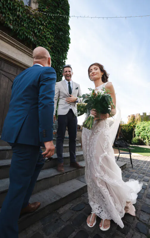 A joyful bride holding a floral bouquet smiles outside a stone manor house while guests laugh and chat on the cobbled courtyard steps.
