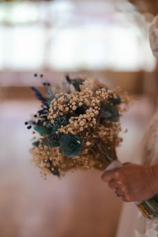 A bride holds a rustic hand-tied bouquet featuring delicate dried gypsophila accented with dusty blue and teal elements against a softly blurred background.