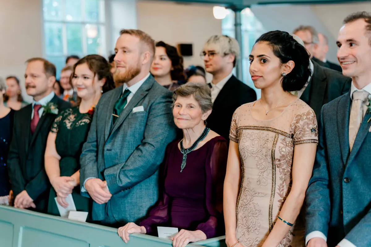 A candid group of well-dressed wedding guests stand together inside a church pew, smiling and observing the ceremony.