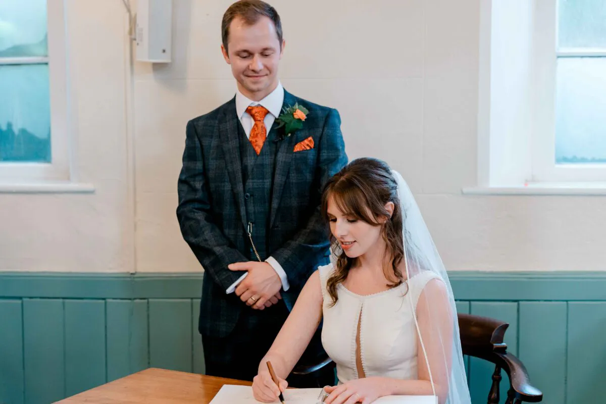 The bride sits at a wooden table signing the register while the groom stands proudly behind her, smiling lovingly down at her during their ceremony.