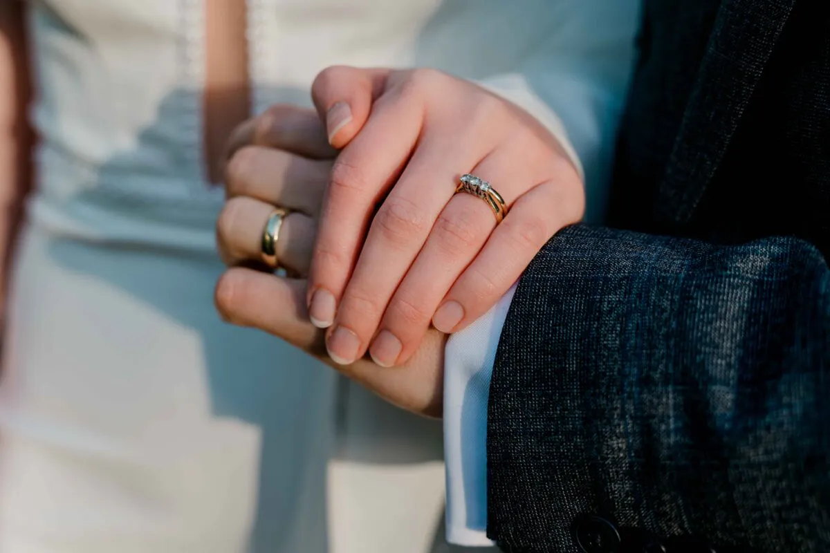 Close-up shot of a newly married couple holding hands, showcasing their wedding bands and a sparkling diamond engagement ring against a charcoal suit and white dress.