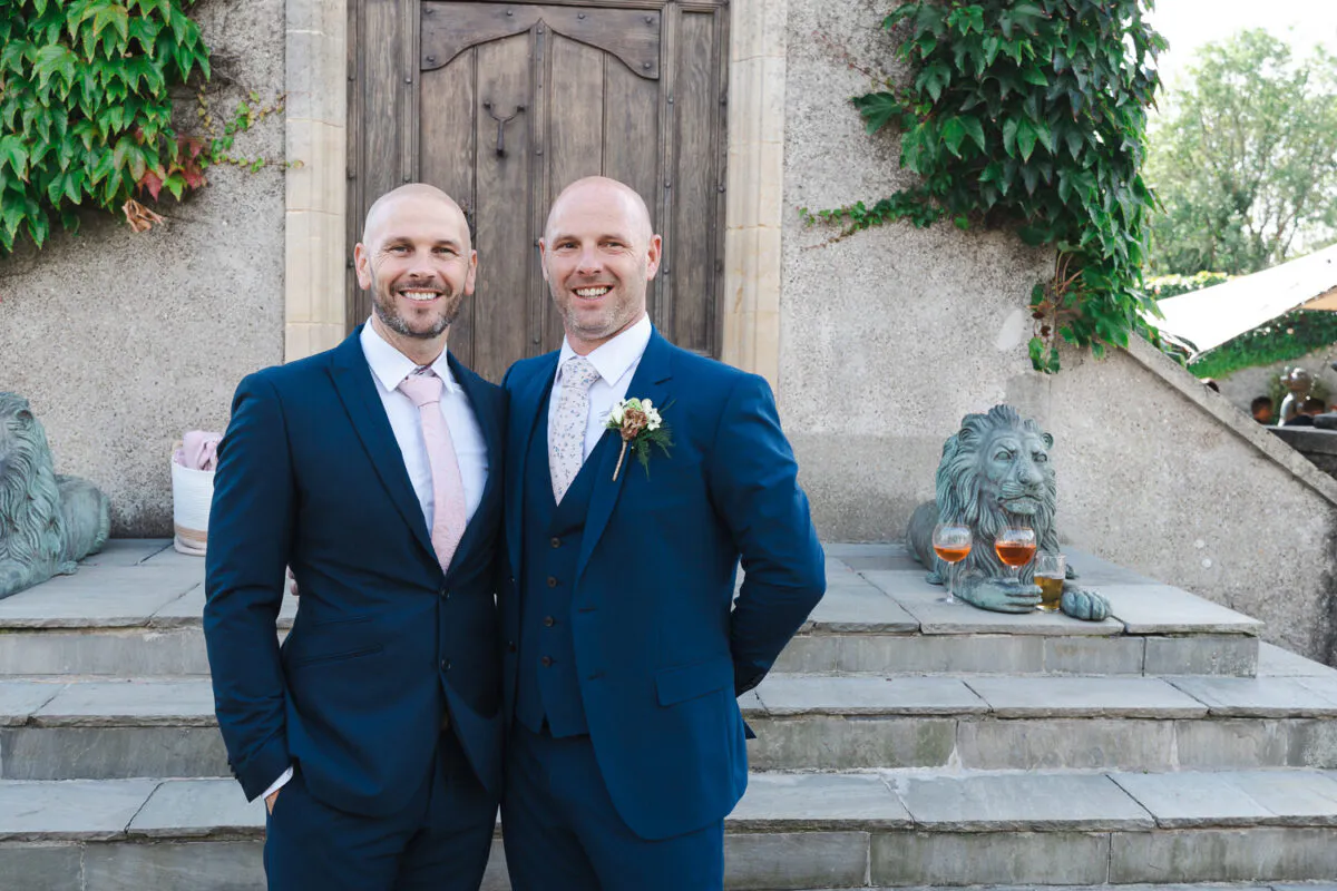 Two smiling grooms in smart navy suits stand side by side on stone steps outside a building framed by climbing ivy.