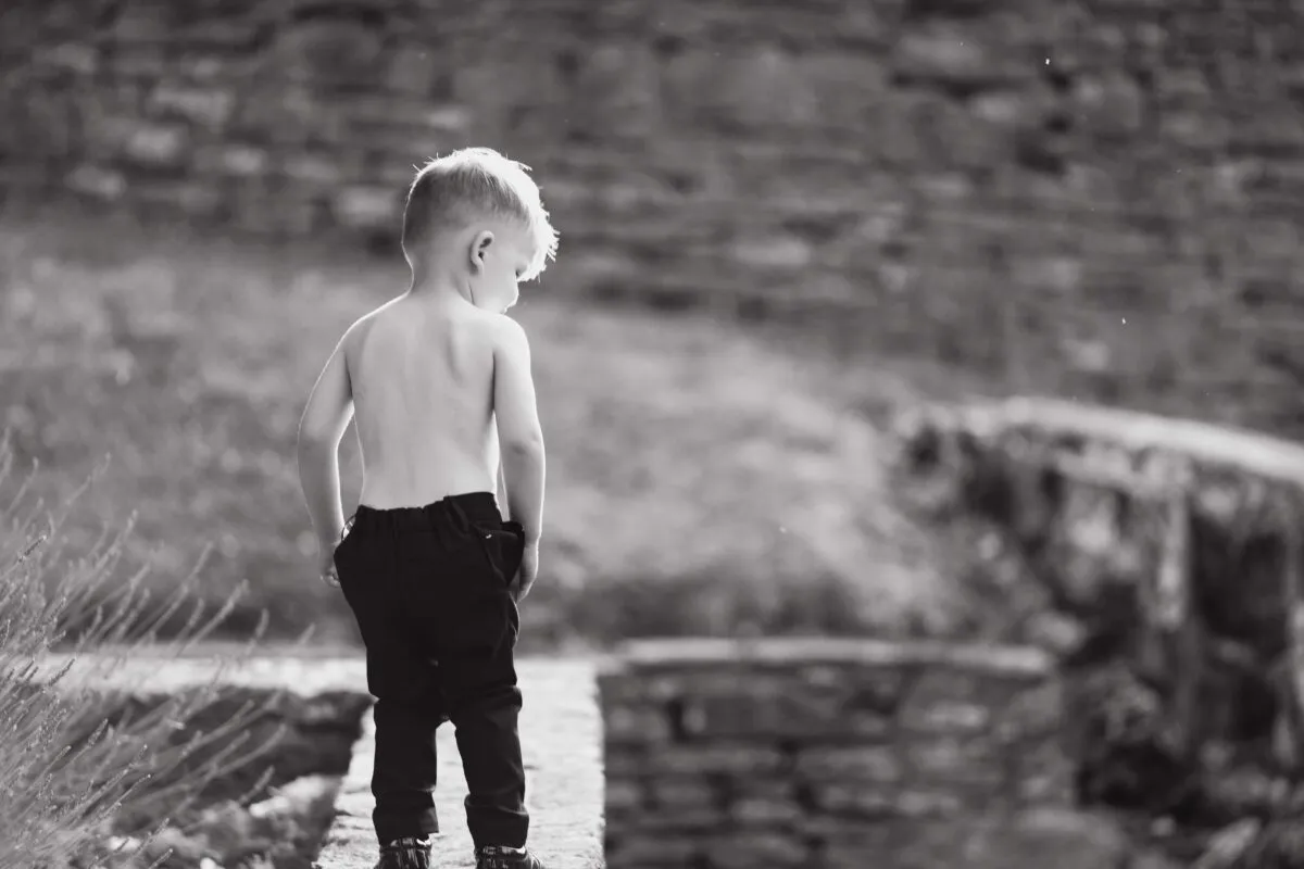 A young page boy stands shirtless from behind, looking down against a rustic stone wall backdrop in a candid black and white shot.