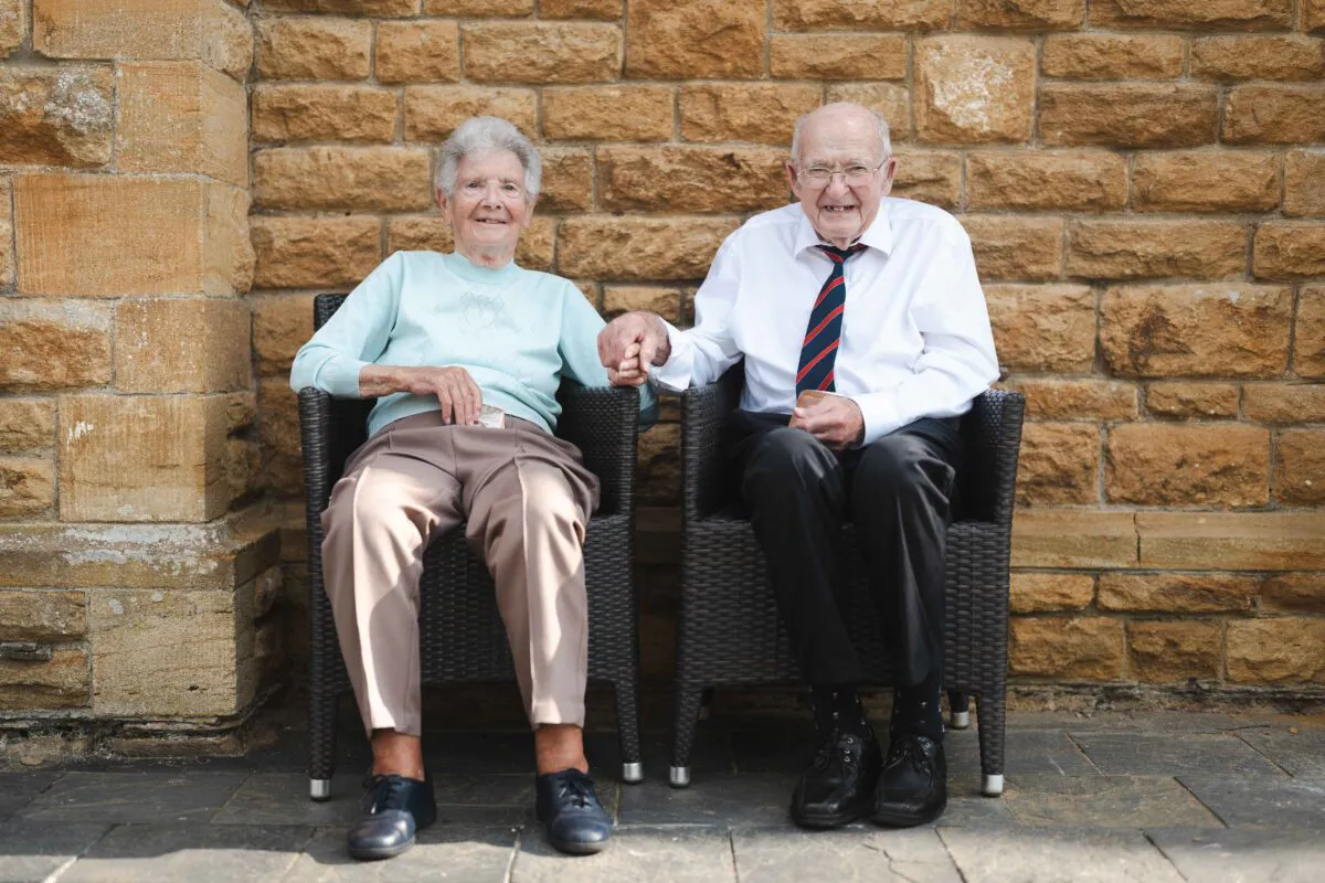 An elderly couple happily sit together on chairs outdoors, holding hands in front of a rustic stone wall.