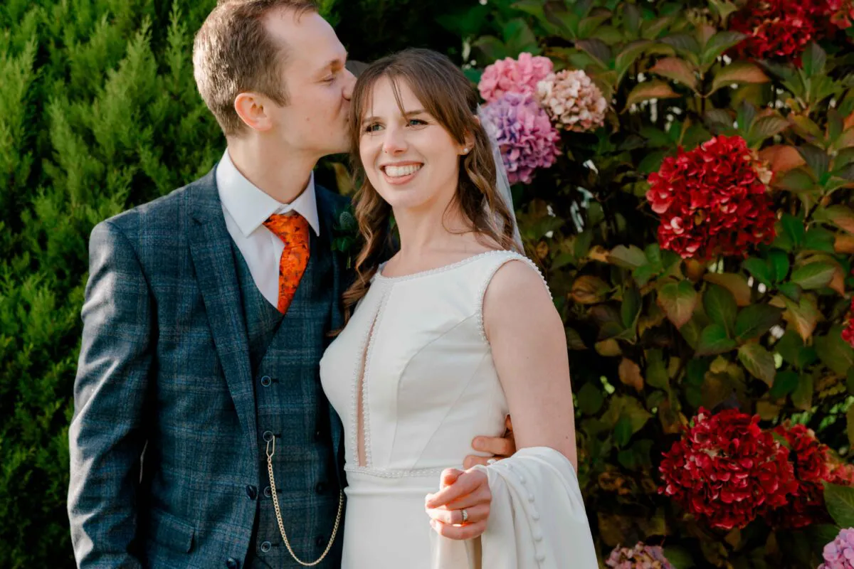 A groom in a blue tweed suit kisses the bride on the forehead as she smiles happily against a backdrop of vibrant hydrangeas.