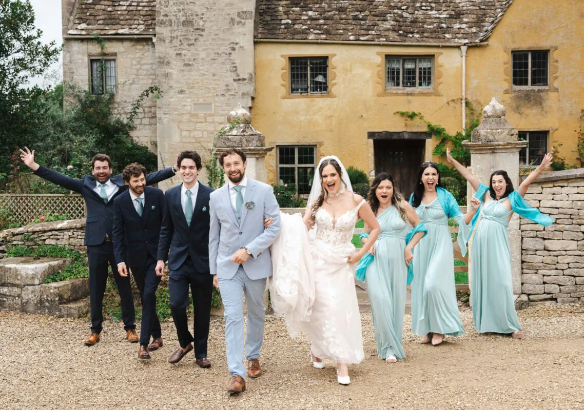 A joyful wedding party of groomsmen in dark and light suits and bridesmaids in flowing sage green dresses walks and laughs together outside a historic stone country house.