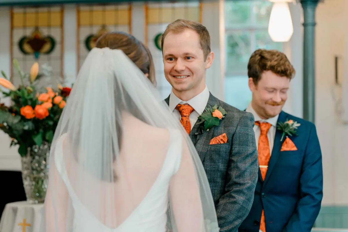 A smiling groom looks lovingly at his bride at the altar during their indoor wedding ceremony, wearing a tweed suit with an orange patterned tie.